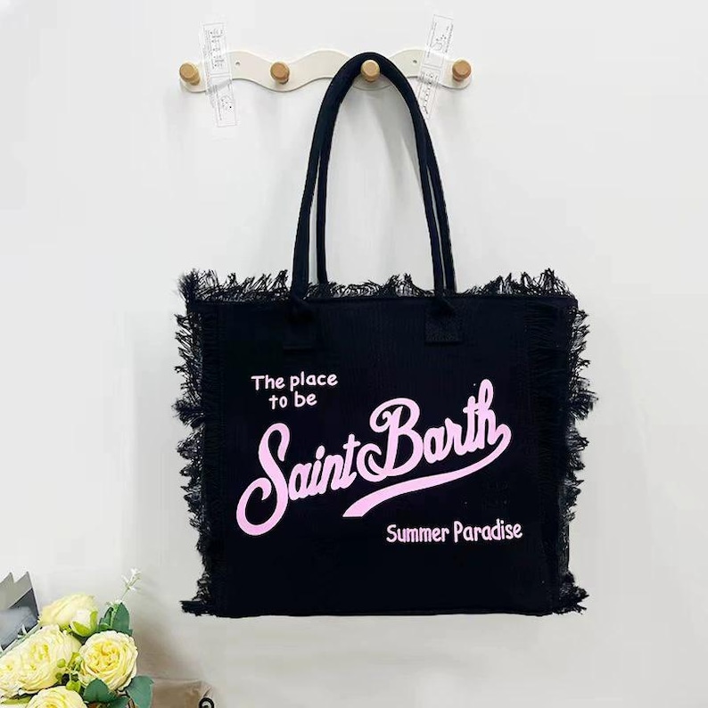 Puede incluir: Bolso tote negro con asas largas y bordes deshilachados. El bolso presenta el texto "The place to be Saint Barth Summer Paradise" en rosa. Cuelga de una pared blanca con un gancho de madera.