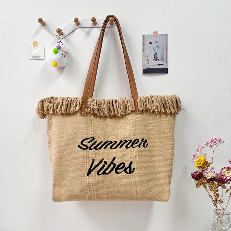 Pu&ograve; includere: Una borsa tote beige con manici in pelle marrone e frange sul bordo superiore. Le parole "Summer Vibes" sono ricamate in nero sul davanti. La borsa &egrave; appesa a una parete bianca.