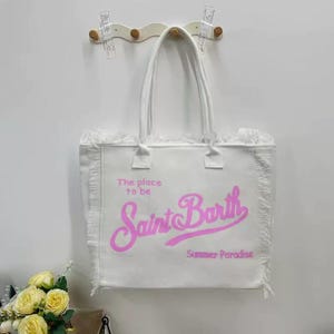 Puede incluir: Bolso tote blanco con ribete de flecos y asas. La bolsa presenta el texto "The place to be Saint Barth Summer Paradise" en letras rosas. La bolsa est&aacute; colgada en una pared.