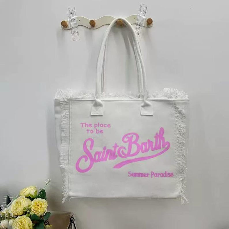 Puede incluir: Bolso tote blanco con ribete de flecos y asas. La bolsa presenta el texto "The place to be Saint Barth Summer Paradise" en letras rosas. La bolsa est&aacute; colgada en una pared.