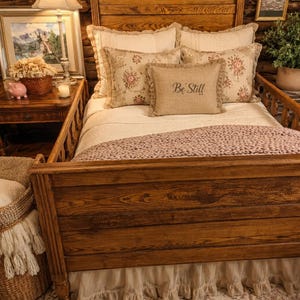 May include: A wooden bed frame with a tall headboard and footboard, featuring a white bedspread, a textured mauve throw blanket, two large white pillows, two floral patterned pillows, and a small rectangular pillow with the text Be Still.