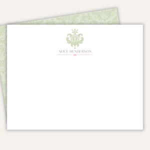 May include: A white rectangular stationery card featuring a sage green damask pattern on the back flap. The front displays a sage green floral crest above the name ALICE HENDERSON in gray text, underlined by a thin pink line with a small bow icon.