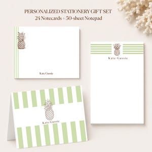 May include: A personalized stationery gift set featuring 24 notecards and a 50-sheet notepad. The items display a brown pineapple illustration, light green stripes, and the name Kate Gussie printed in brown text on white paper surfaces.