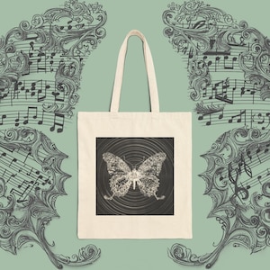 May include: A beige canvas tote bag featuring a centred square graphic of a butterfly composed of musical notes set against a background of concentric circular lines. The bag has two long shoulder straps and a simple rectangular construction.