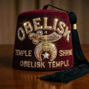 以下が含まれることがあります： 金色の刺繍と輝くラインストーンが施された深紅のベルベット製フェズ帽。テキストには「OBELISK」「TEMPLE」「SHRINE」「OBELISK TEMPLE」と記されている。中央のエンブレムにはシミター、三日月、ファラオの顔が描かれ、長い黒いタッセルが付いている。