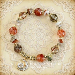 May include: A beaded bracelet featuring a mix of orange, brown, and clear gemstones, accented with gold-colored metal beads and a crescent moon charm. The bracelet is displayed on a cream-colored lace doily.