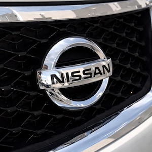 Puede incluir: Primer plano de la parrilla de un coche con el logotipo de Nissan. El logotipo es un emblema plateado y circular con el nombre de la marca en letras negras en negrita. La parrilla tiene un patrón de panal de abeja negro con un borde cromado brillante.