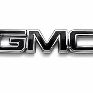 May include: A chrome and black GMC emblem. The letters G, M, and C are in a bold, sans-serif font. The emblem has a shiny chrome border and a black interior, creating a three-dimensional effect. The emblem is on a white background.