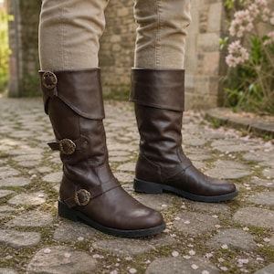 May include: Brown leather boots with buckle detailing. The boots are mid-calf length and have a flat sole. The buckles are gold-toned. The boots are worn with beige trousers. The boots' height is approximately 30 cm.