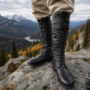 May include: Black leather knee-high boots with lace-up fronts and buckle straps. The boots are worn outdoors on a rocky surface, with a scenic mountain and forest backdrop.