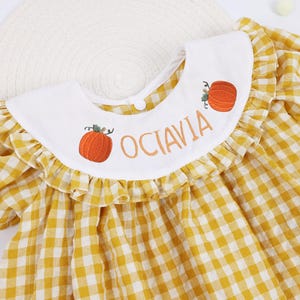 May include: A yellow and white gingham dress featuring a white Peter Pan collar with a ruffled edge. The collar is embroidered with two orange pumpkins and the name OCTAVIA in orange lettering. The dress has short puff sleeves and a button closure.