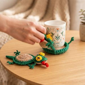 May include: Two handmade crochet coasters shaped like green and tan lizards with yellow eyes, black cross-stitched pupils, and red tongues. One coaster lies flat on the surface, while the other is wrapped around a white ceramic mug with floral art.