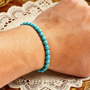 Könnte beinhalten: Ein türkisfarbenes Perlenarmband. Das Armband besteht aus runden, hellblauen Perlen mit marmoriertem Aussehen. Das Armband wird am Handgelenk getragen. Der Text "TINY GLEAM JEWELRY" ist unten sichtbar.