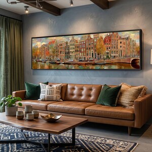 May include: A panoramic wall art print featuring a row of narrow, multi-story canal houses in Amsterdam. The painting displays warm autumn tones, reflections on the water, and a stone bridge, all framed in a thin black border on a gray wall.