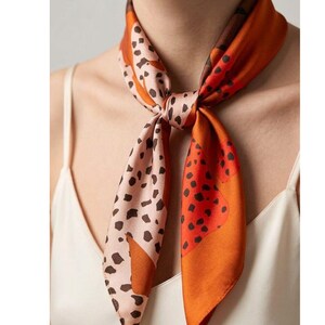 May include: A patterned silk scarf tied in a knot. The scarf displays a design of brown spots on a pale pink backdrop, alongside orange and brown panels. The scarf is worn around the neck, offering a fashionable accessory.