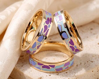 Puede incluir: Tres anillos de metal en tono dorado con un patrón de incrustaciones de esmalte de colores. El diseño consiste en formas irregulares tipo mosaico en tonos morado, azul claro y lavanda, sobre la superficie dorada pulida del anillo.