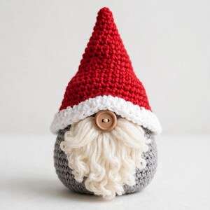 May include: A crochet gnome figure featuring a tall, pointed red hat with a white brim. The gnome has a round, light brown wooden button nose centered above a thick, textured, off-white yarn beard. The base of the gnome is gray crochet.
