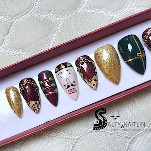 May include: A set of press-on nails in a red box. The nails feature various designs, including gold glitter, a nutcracker face, and dark green with gold accents. Some nails have gold leaf and gem embellishments.