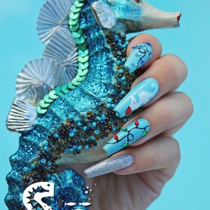 Peut inclure: Une main avec un vernis à ongles bleu et argenté pailleté. Les ongles sont décorés d'un motif de cheval marin et de lumières de Noël. La main tient un ornement de cheval marin bleu et vert. L'ornement porte le texte "Salty Kaitlin LLC Cape Coral, Florida".