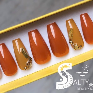 May include: Set of ten false nails with a fall theme. The nails are in shades of brown and gold with gold leaf accents and glitter. The nails are displayed in a yellow box with the text "Salty Kaitlin Beachy Nails & More".