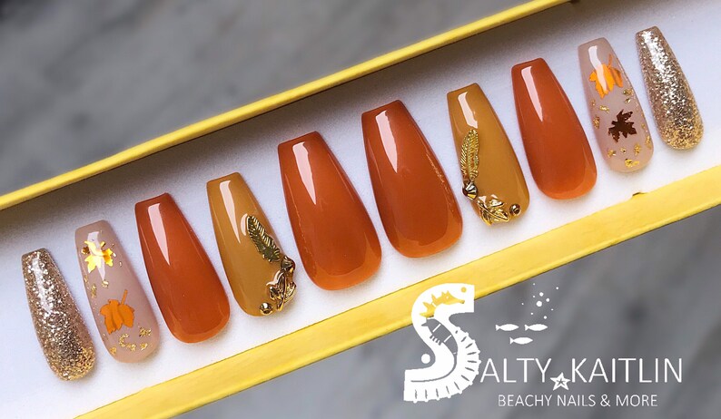 May include: Set of ten false nails with a fall theme. The nails are in shades of brown and gold with gold leaf accents and glitter. The nails are displayed in a yellow box with the text "Salty Kaitlin Beachy Nails & More".