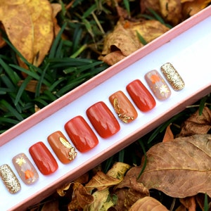 May include: A set of eight false nails with a fall-themed design. The nails are in a variety of shades of orange and brown, with gold leaf accents and glitter. The nails are displayed in a pink box.