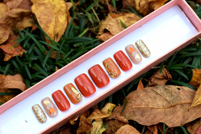 May include: A set of eight false nails with a fall-themed design. The nails are in a variety of shades of orange and brown, with gold leaf accents and glitter. The nails are displayed in a pink box.