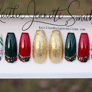 Puede incluir: Conjunto de diez u&ntilde;as postizas con tem&aacute;tica navide&ntilde;a. Las u&ntilde;as est&aacute;n decoradas con purpurina dorada, pintura verde, roja y blanca, y peque&ntilde;os adornos. Las u&ntilde;as est&aacute;n dispuestas en fila sobre una superficie blanca. kailtlinsdesignernailsspa.com