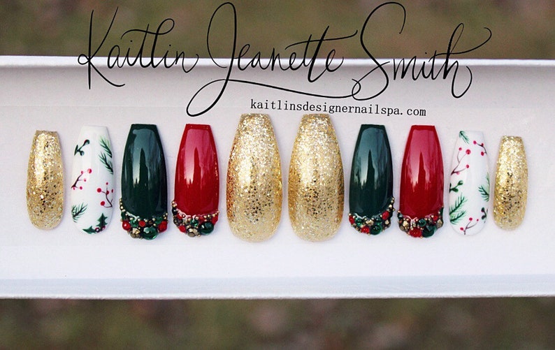 Puede incluir: Conjunto de diez u&ntilde;as postizas con tem&aacute;tica navide&ntilde;a. Las u&ntilde;as est&aacute;n decoradas con purpurina dorada, pintura verde, roja y blanca, y peque&ntilde;os adornos. Las u&ntilde;as est&aacute;n dispuestas en fila sobre una superficie blanca. kailtlinsdesignernailsspa.com