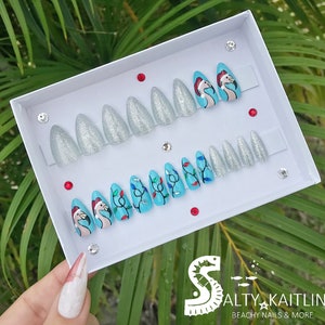 Peut inclure: Un ensemble de 10 faux ongles sur le thème de Noël. Les ongles sont en paillettes argentées et bleu turquoise avec un motif de flamant rose et des lumières de Noël.