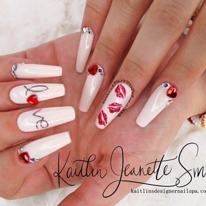 Puede incluir: U&ntilde;as acr&iacute;licas largas, blancas, en forma de ata&uacute;d con corazones rojos y la palabra "love" escrita en ellas. Algunas u&ntilde;as tienen diamantes de imitaci&oacute;n y brillo. Las u&ntilde;as est&aacute;n decoradas con labios rojos brillantes y el texto "Kaitlin Jeanette Smith" y "kaitlinsdesignernailsspa.com".