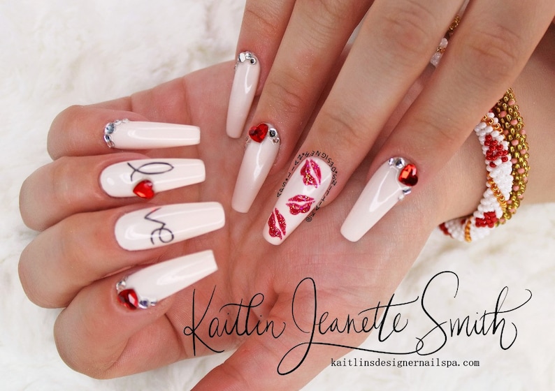 Puede incluir: U&ntilde;as acr&iacute;licas largas, blancas, en forma de ata&uacute;d con corazones rojos y la palabra "love" escrita en ellas. Algunas u&ntilde;as tienen diamantes de imitaci&oacute;n y brillo. Las u&ntilde;as est&aacute;n decoradas con labios rojos brillantes y el texto "Kaitlin Jeanette Smith" y "kaitlinsdesignernailsspa.com".