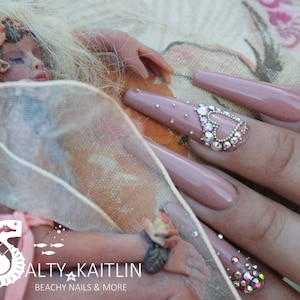May include: Long, nude-colored acrylic nails with rhinestones and a heart-shaped design. The nails are decorated with small, clear rhinestones and larger, iridescent rhinestones. The heart is outlined with rhinestones and filled with smaller rhinestones.