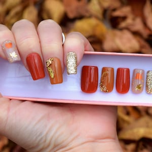May include: Set of ten false nails with a fall theme. The nails are in various shades of orange and brown with gold leaf accents and glitter.