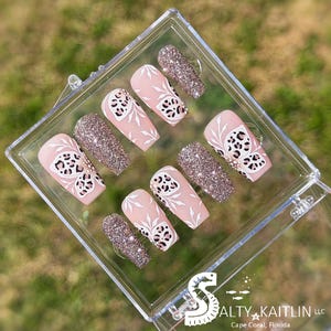 May include: A set of false nails in a clear case. The nails have a light pink base with white floral designs and black leopard print accents. Some nails are covered in glitter.
