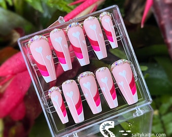 May include: A set of ten pink and white coffin-shaped press-on nails with a white heart design and pink and red stripes. The nails are decorated with rhinestones and pearls.