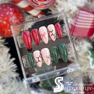 Peut inclure: Une boîte en plastique transparente contenant dix faux ongles. Les ongles sont décorés de paillettes rouges et vertes, et certains ont un motif de nœud rouge et de houx vert avec des baies rouges. La boîte porte le logo de Salty Kaitlin LLC, Cape Coral, Floride.