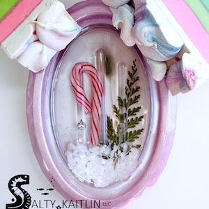 May include: A pink oval-shaped resin soap dish with a candy cane, greenery, and faux snow inside. The dish is decorated with white and blue swirls and a pink and green bar of soap. The text "SALTY KAITLIN LLC Cape Coral, Florida" is at the bottom.