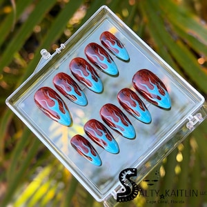 May include: A set of ten artificial nails in a clear plastic case. Each nail is oval-shaped with a glossy finish. The design features a brown base with blue flame-like accents. The nails are arranged in a clear plastic case. The text "SALTY KAITLIN LLC" is visible.