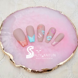 May include: Set of five nude-colored false nails with a pink and blue glitter accent on the tips. The nails are decorated with pink and white lips. The nails are displayed on a pink and gold agate-style tray with the text "Salty Kaitlin Beachy Nails & More".