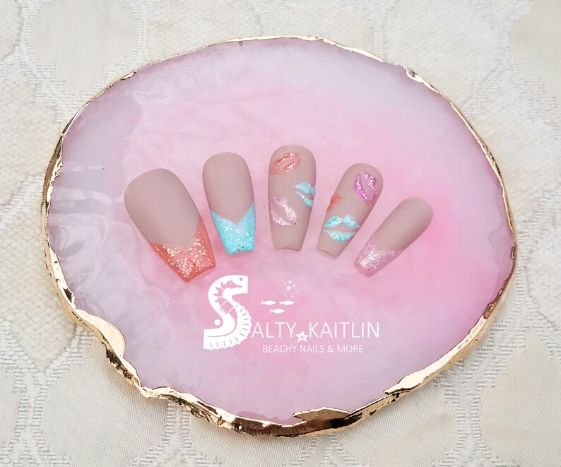 May include: Set of five nude-colored false nails with a pink and blue glitter accent on the tips. The nails are decorated with pink and white lips. The nails are displayed on a pink and gold agate-style tray with the text "Salty Kaitlin Beachy Nails & More".