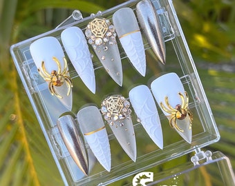 May include: A set of ten false nails in a clear plastic box. The nails are white with a spiderweb design and gold spider accents. Some nails have a silver glitter finish. The box has a logo for Salty Kaitlin LLC, Cape Coral, Florida.