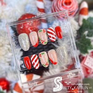 Peut inclure: Un ensemble de six faux ongles sur le thème de Noël. Les ongles sont décorés de rayures de cannes en sucre rouge et blanc, de feuilles de houx et de baies rouges. Deux des ongles sont en paillettes argentées.