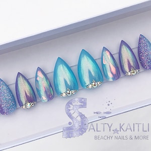 May include: Set of ten iridescent blue and purple stiletto false nails with glitter and rhinestones. The nails are arranged in a box with the text "Salty Kaitlin Beachy Nails & More".