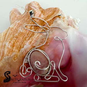 May include: A silver wire pendant in the shape of a rabbit, featuring an oval, pink gemstone center. The pendant is displayed on a seashell. The text "Salty Kaitlin" is visible in the lower left corner.