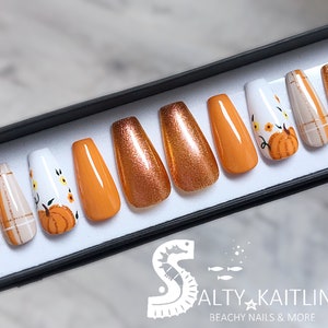 Peut inclure: Un ensemble de dix faux ongles dans une bo&icirc;te noire. Les ongles sont en forme de cercueil et pr&eacute;sentent une vari&eacute;t&eacute; de motifs. Certains ongles sont orange, d'autres sont bruns avec des paillettes et d'autres sont blancs avec un motif de citrouille et de fleurs. La bo&icirc;te porte le texte "Salty Kaitlin Beachy Nails & More" en bas.
