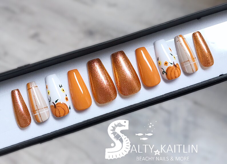 Peut inclure: Un ensemble de dix faux ongles dans une bo&icirc;te noire. Les ongles sont en forme de cercueil et pr&eacute;sentent une vari&eacute;t&eacute; de motifs. Certains ongles sont orange, d'autres sont bruns avec des paillettes et d'autres sont blancs avec un motif de citrouille et de fleurs. La bo&icirc;te porte le texte "Salty Kaitlin Beachy Nails & More" en bas.