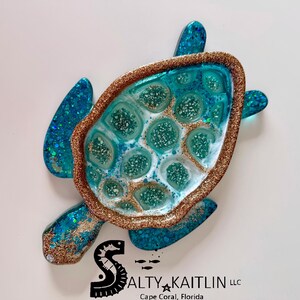 May include: A handcrafted sea turtle wall art piece. The turtle features a turquoise and gold glitter design with a textured, resin-like finish. The edges are outlined in gold glitter. The piece is a decorative item.