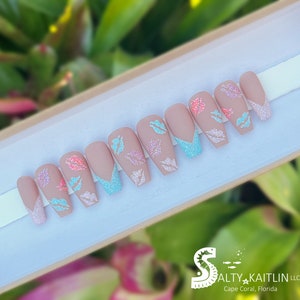 May include: Set of ten nude-colored press-on nails with a light blue glitter accent and pink and blue glitter butterfly designs. The nails are displayed in a white box with the text "Salty Kaitlin LLC Cape Coral, Florida".