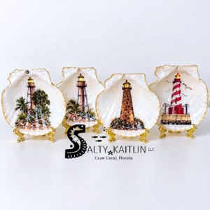 May include: Four decorative seashell dishes with gold-painted edges, each resting on a small gold stand. The shells feature detailed lighthouse illustrations, including palm trees, rocky bases, and a red-and-white striped tower. Text reads SALTY KAITLIN LLC.